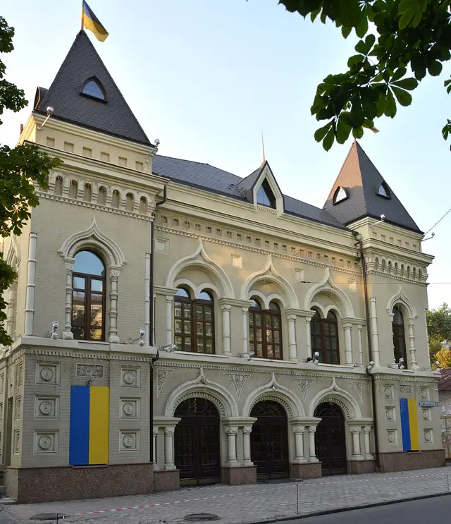 Літній фасад історичної будівлі на Володимира Вернадського, 23 із зеленим листям, прапорами України та адресною табличкою