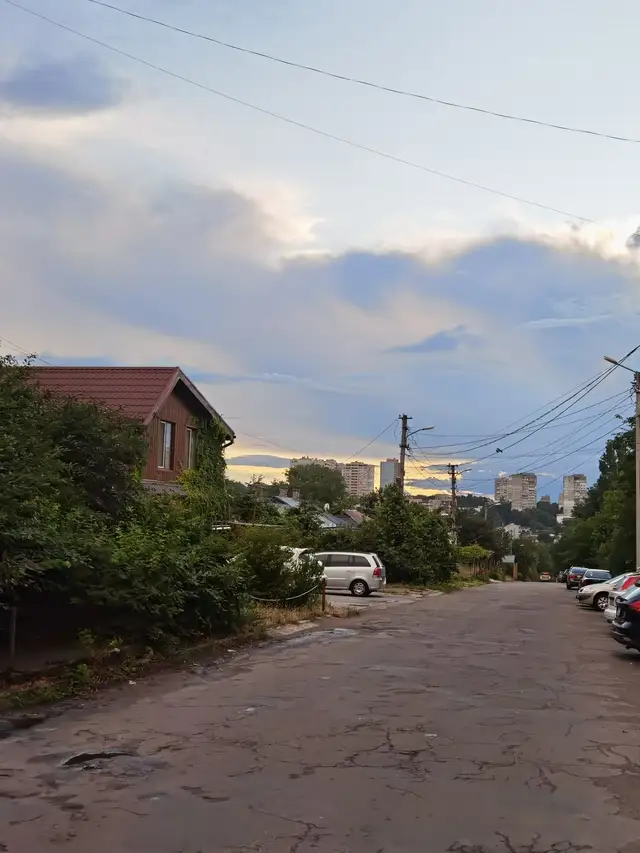 Літня вулиця Леоніда Стромцова з зеленими садибами, спуском до багатоповерхової забудови та вечірнім небом