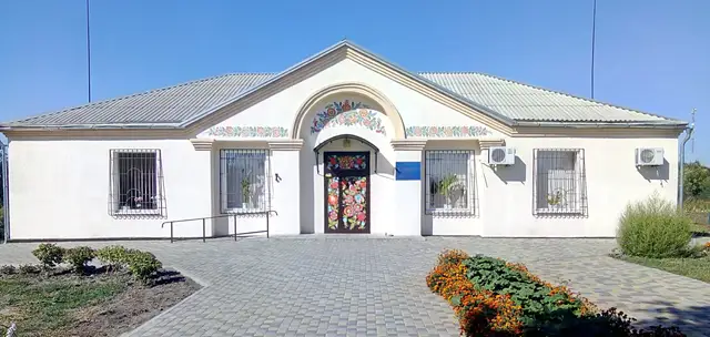Світлий одноповерховий фасад Петриківського музею з петриківським квітковим розписом над входом, ґратованими вікнами та квітником