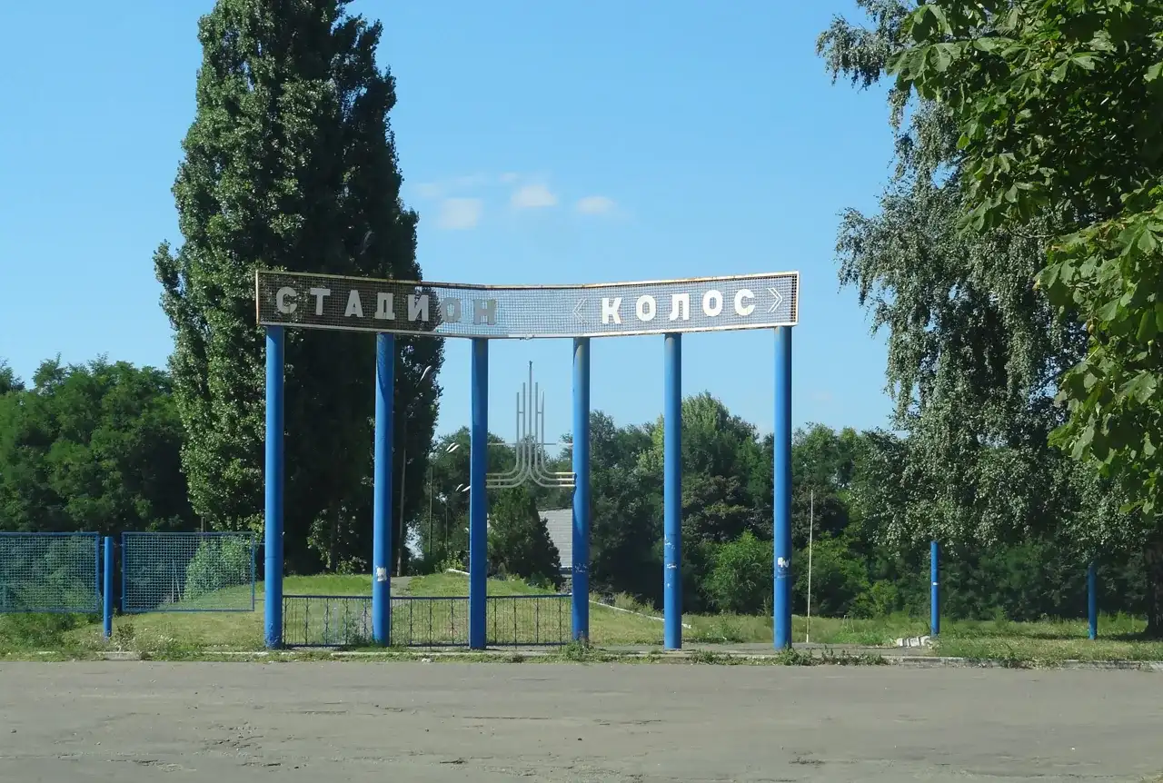 Вхід до стадіону з написом «СТАДИОН КОЛОС» між деревами у Солоному