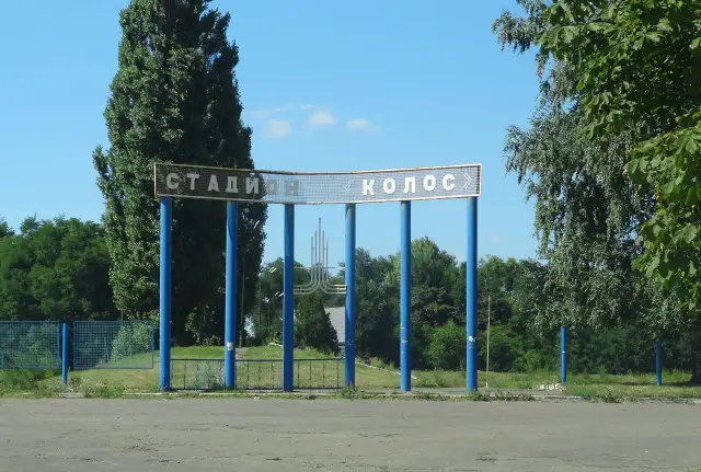 Вхід до стадіону з написом «СТАДИОН КОЛОС» між деревами у Солоному
