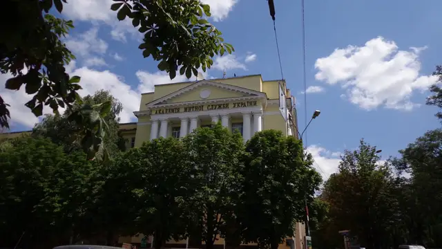 Літній фасад навчального корпусу УМСФ серед зелених дерев з історичним написом митної академії